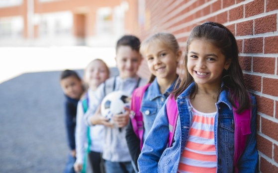 Five smiling kids with backpacks standing next to red brick wall